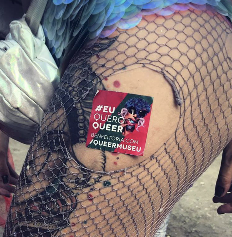 Foto de um adesivo escrito #EuQueroQueer da campanha da exposição QueerMuseu colado na coxa de uma foliã do carnaval carioca, com uma meia arrastão rasgada e uma tatuagem de colombina.